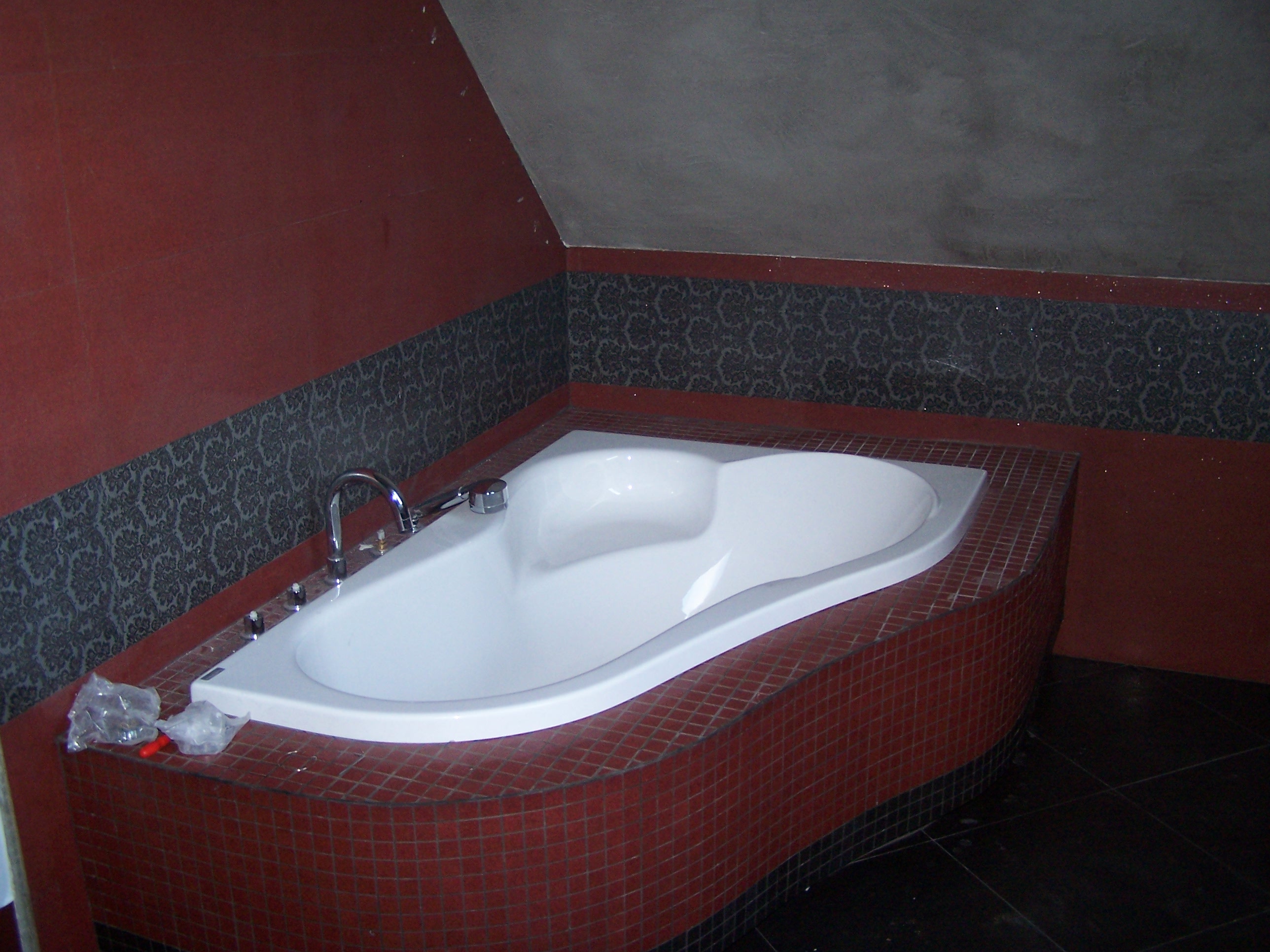 Narożna wanna z hydromasażem obudowana czerwoną mozaiką, widoczne elementy instalacji wodnej, fragment ściany w kolorze ceglastym i dekoracyjny pas tapety w łazience na poddaszu.